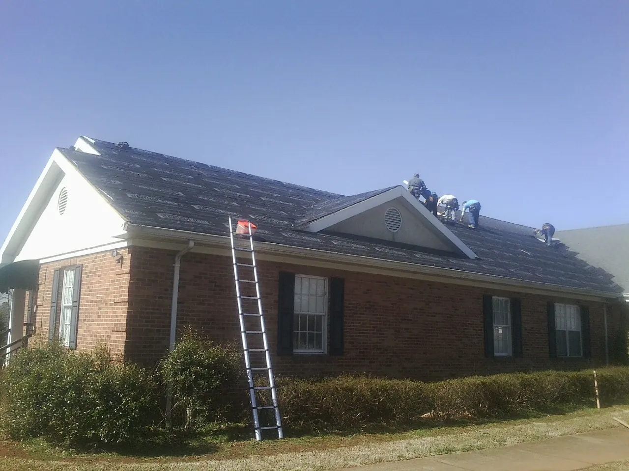 Crew tearing shingles off a brick ranch roof with a ladder set — Emergency Roof Repair work by our crew in Mc Ewen