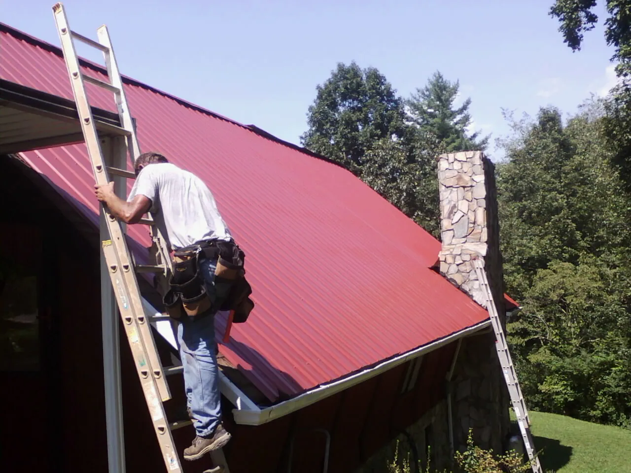 Roofer on a ladder beside a red metal roof and stone chimney — Soffit & Fascia Repair by Roofing Brothers in Mc Ewen